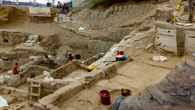 Archaeologists excavating a large Bronze Age site with ancient walls and layered soil structures. 
