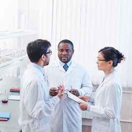 Three scientists in lab coats discussing their research in a lab with lab glassware on the shelf behind them. 