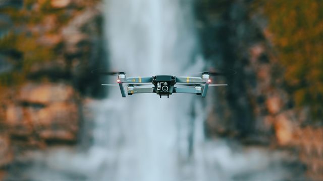 A silver drone hovering in front of a waterfall and stream. 