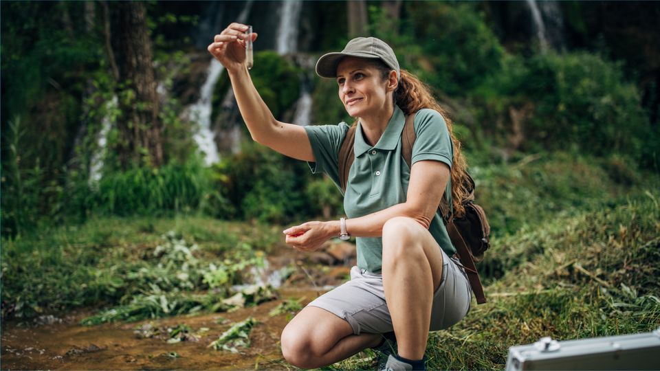 A female environmental scientist kneeling by a stream in a forest, examining a water sample in a test tube, with field equipment beside her, conducting environmental analysis in a natural setting.