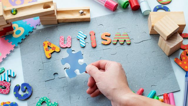 Colorful puzzle pieces and toys arranged around the word "Autism" with a hand placing a piece. 